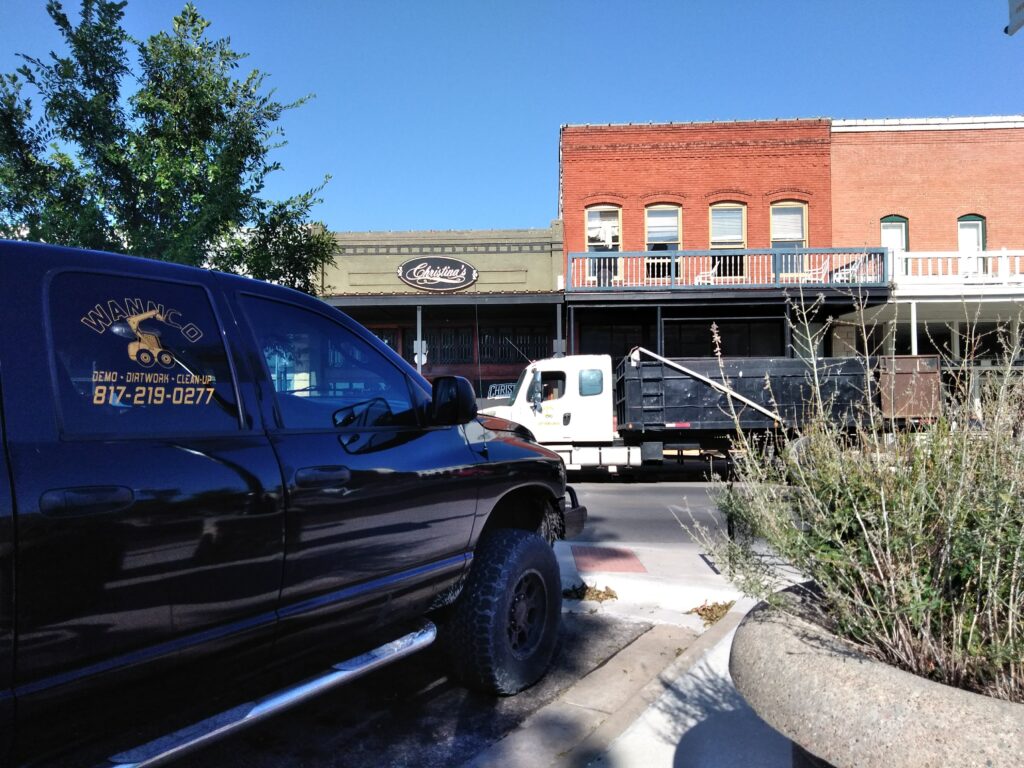 View of a Wannco Services demolition contractor truck parked in front of a historic building in the Granbury Square in Granbury, Texas. The building has a dump truck in front for demolition scraps.