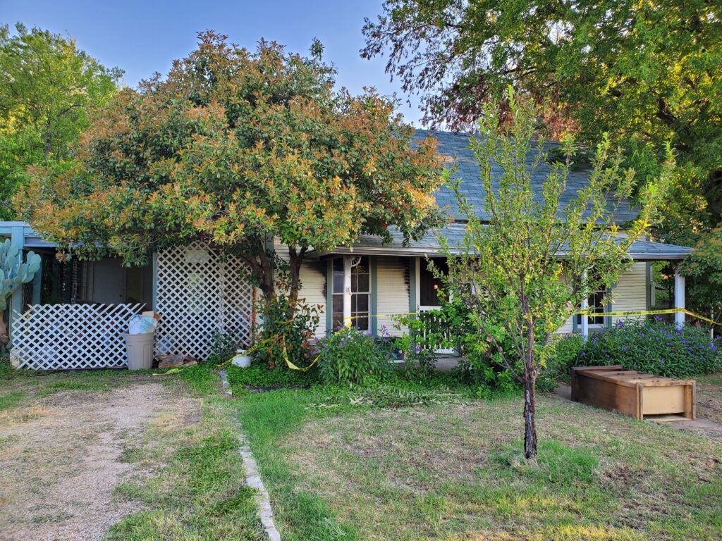 An old house with vegetation in the front including a tree and rose bush being prepped for demolition