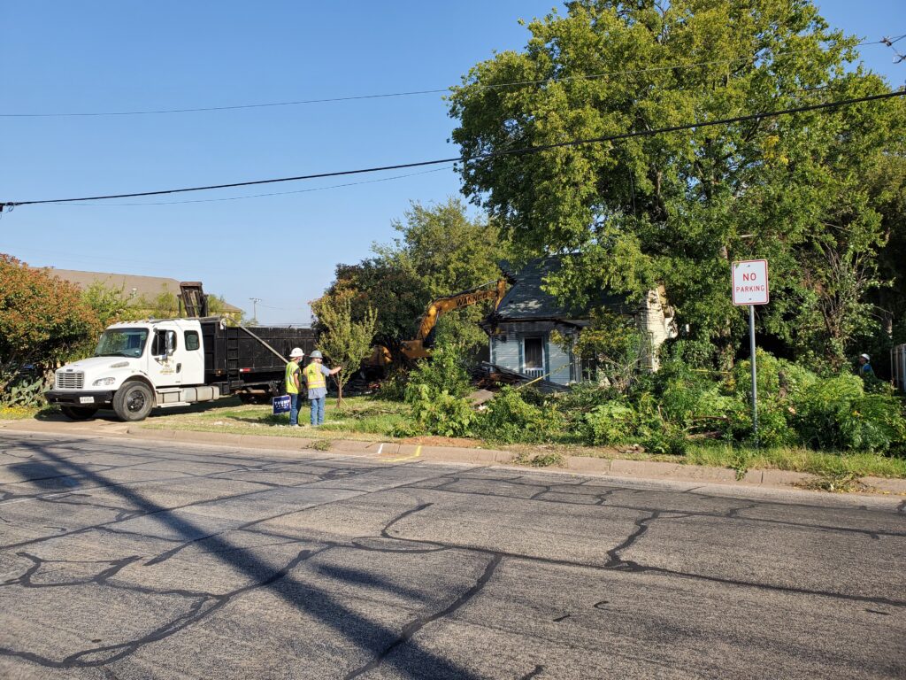 A street view of an old house with vegetation in the front including a tree and rose bush being prepped for demolition